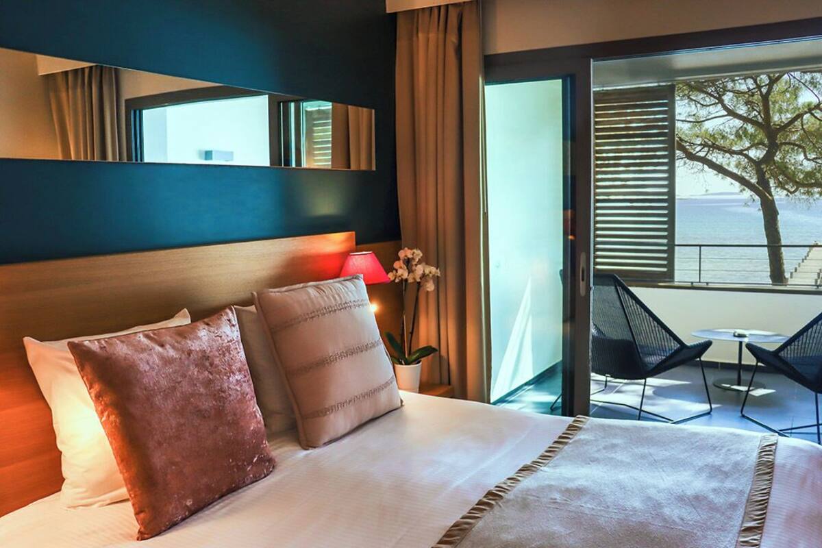 A hotel room with a bed, a large window, a balcony with chairs, and a view of the sea.