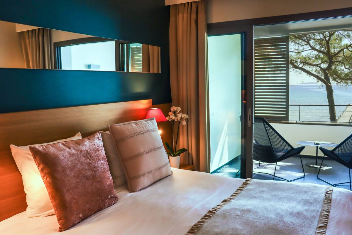 A hotel room with a large bed, a nightstand with a lamp, and a view of the sea through a sliding door.