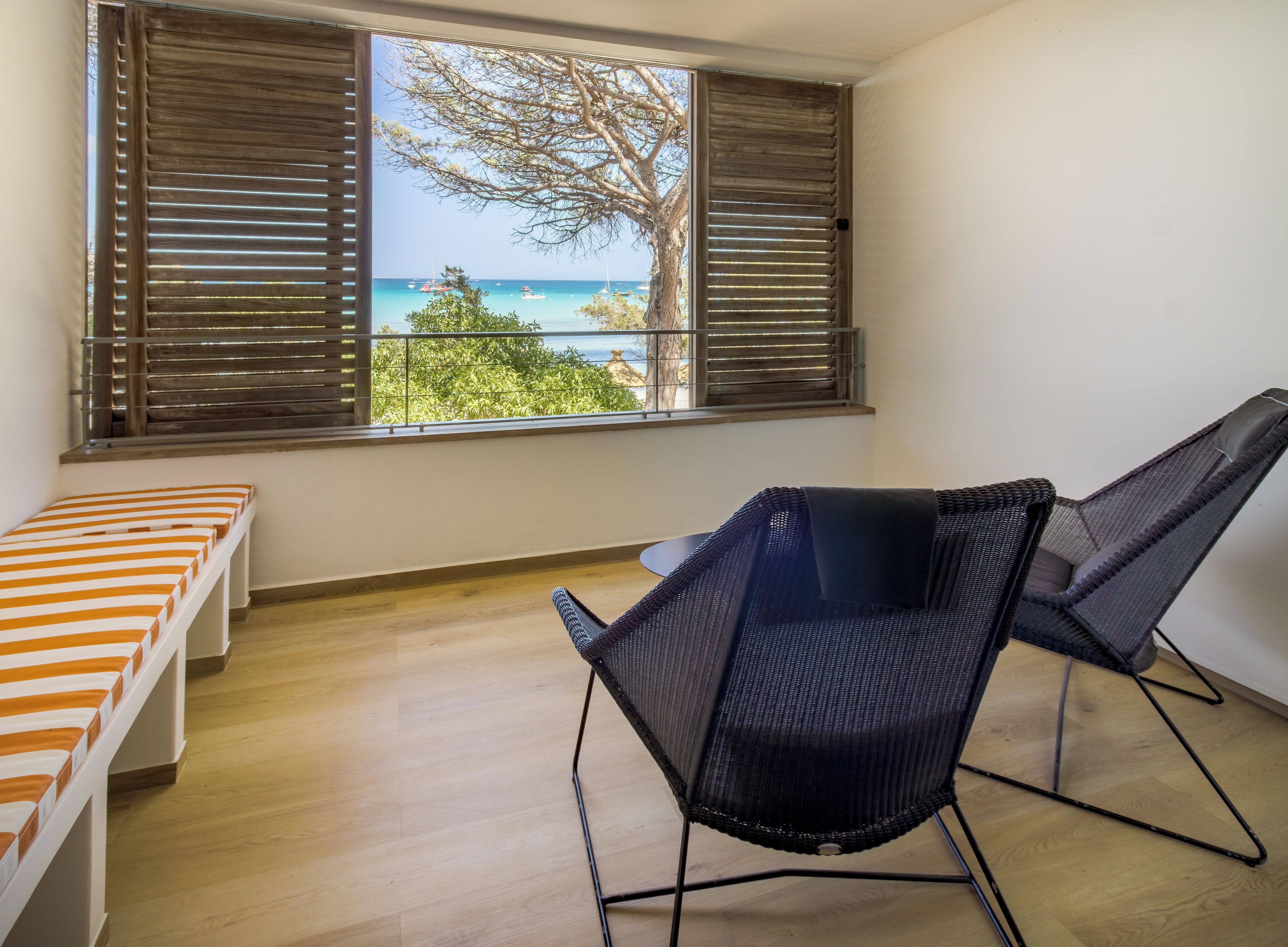 A room with a wooden floor, a bed with striped bedding, a chair, and a window with blinds overlooking a view of the sea and trees.