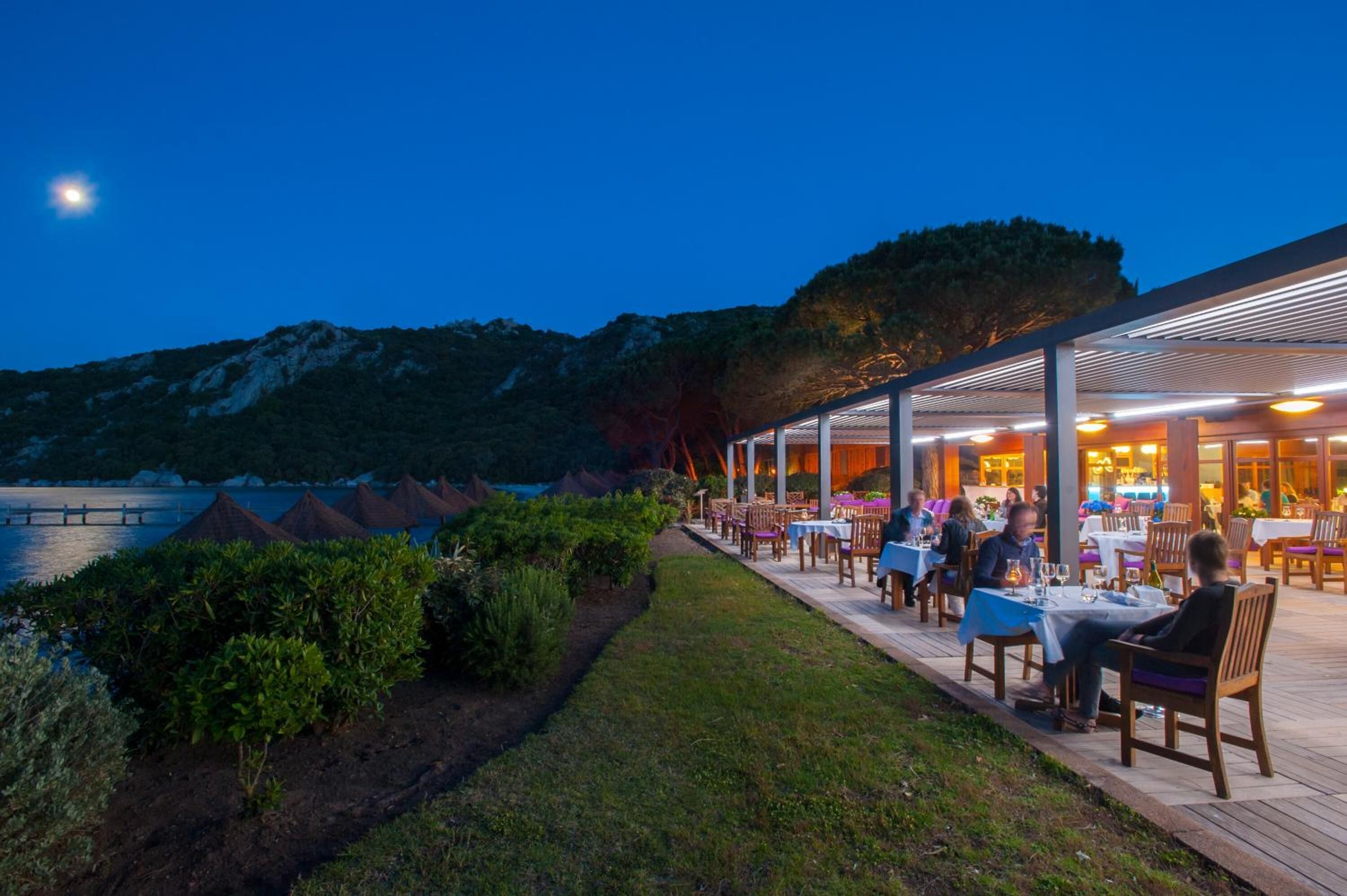 An outdoor dining area with tables and chairs, overlooking a body of water and hills.