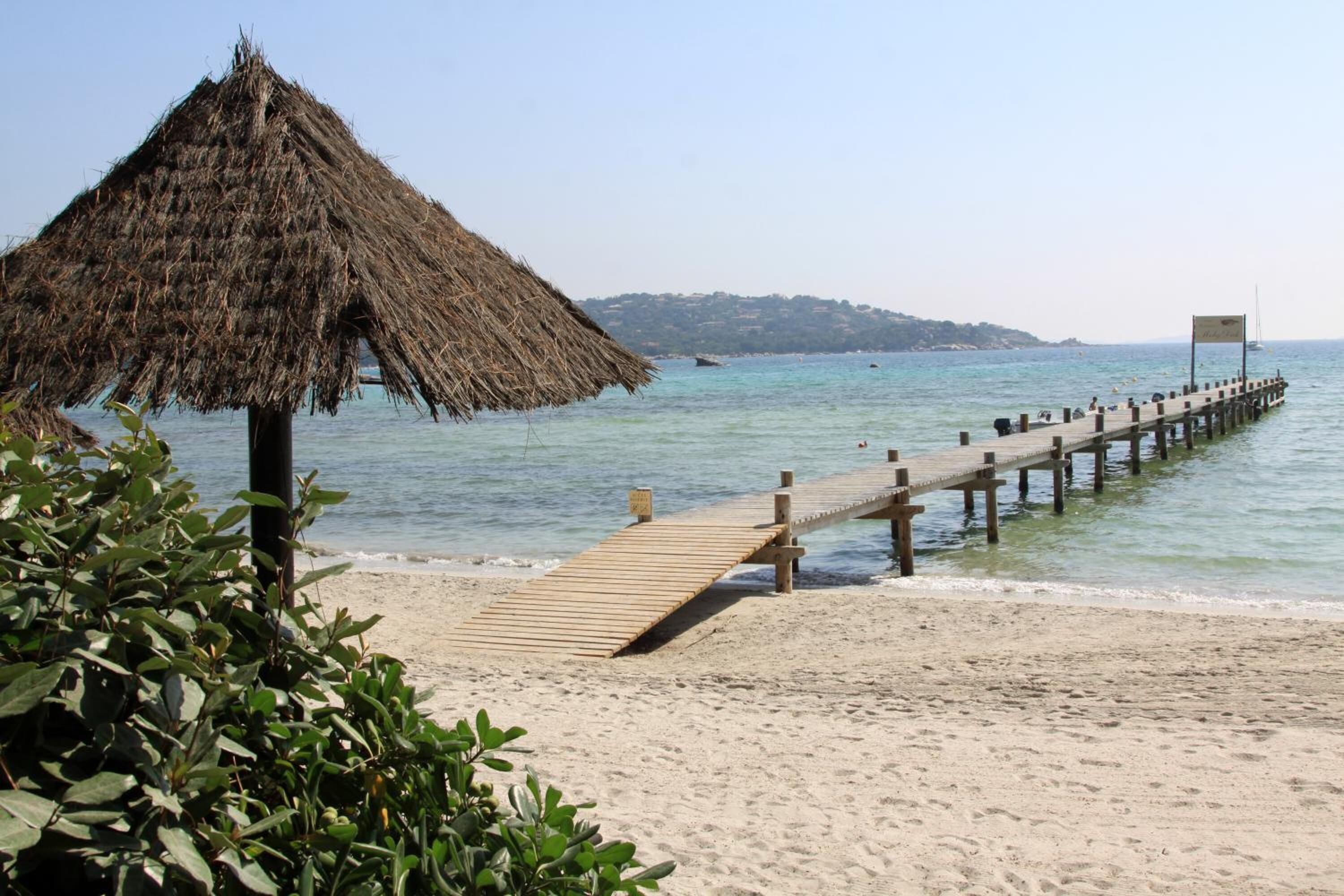 A beach with a wooden pier extending into calm waters, a thatched-roof structure, and a distant landmass.