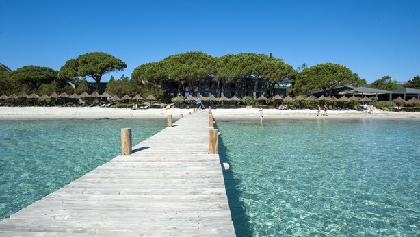 Eine Holzsteige führt zu einem Strand mit klarem Wasser, Sonnenschirmen und Bäumen.