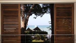 Blick durch ein schmales Holzfenster mit Lamellen auf einen Strand mit Hütten mit Reetdach und einen Baum.