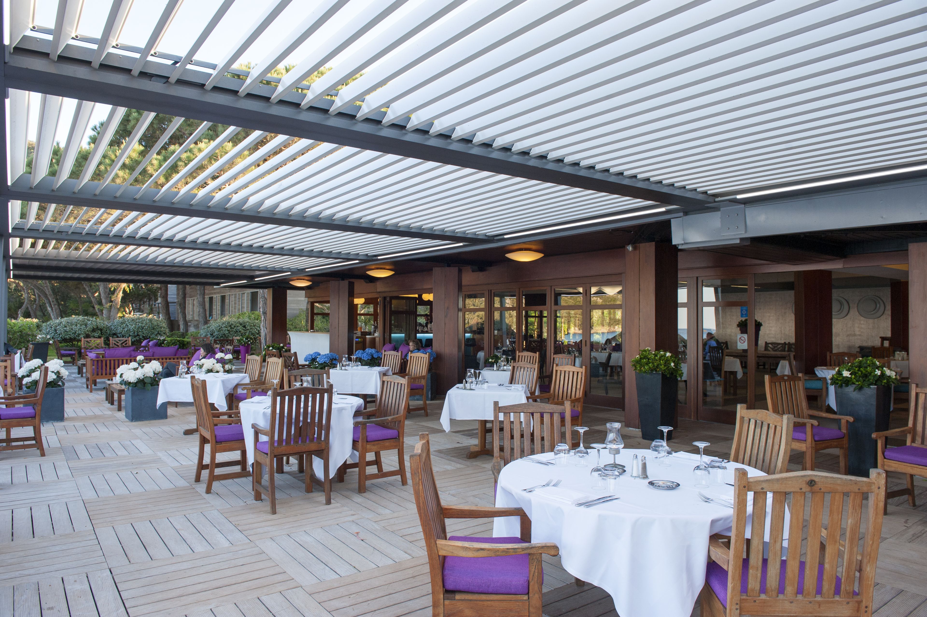An outdoor dining area with tables and chairs, covered by a roof, and surrounded by greenery.