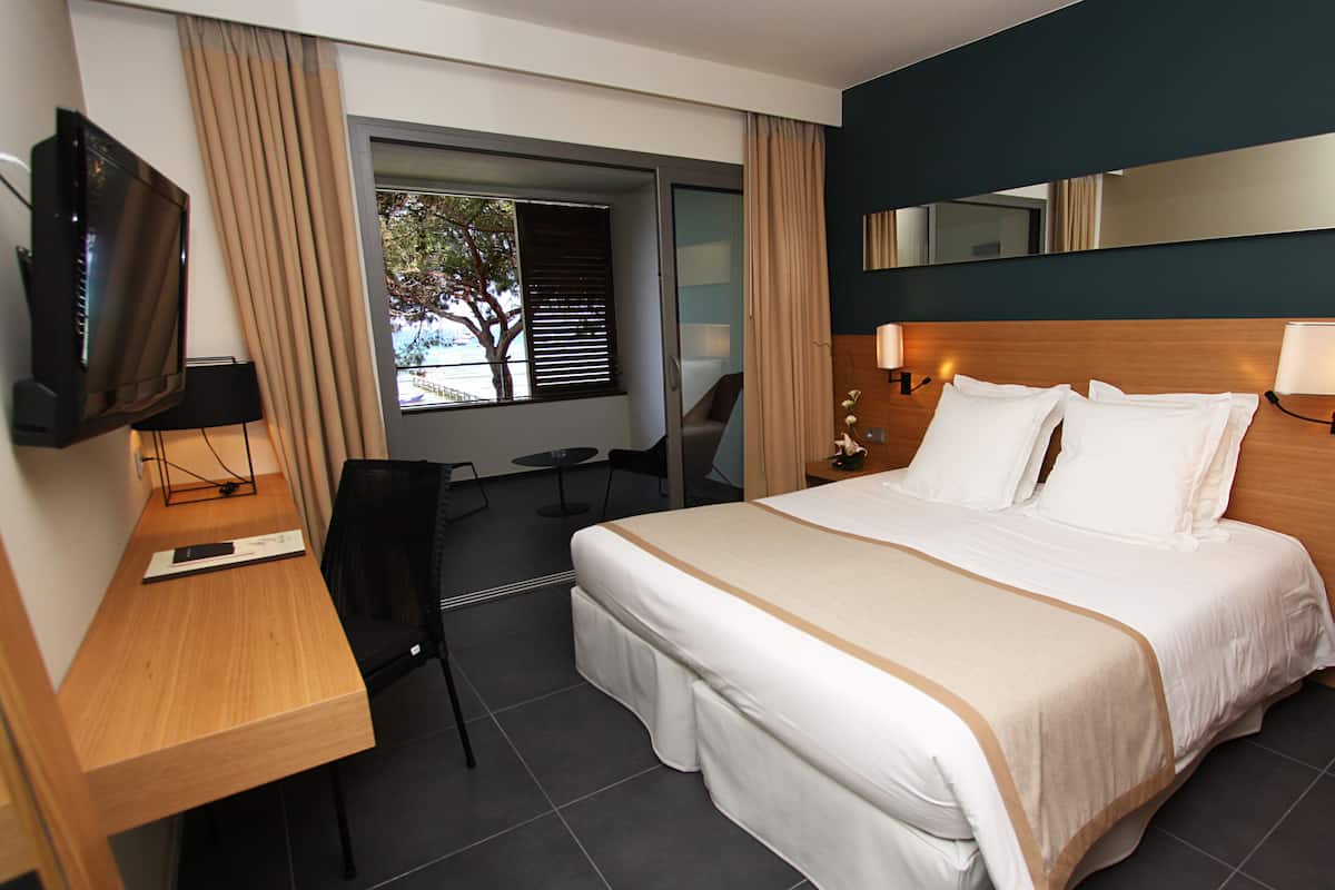 A hotel room with a large bed, a desk with a computer, a TV, a chair, and a window with curtains.
