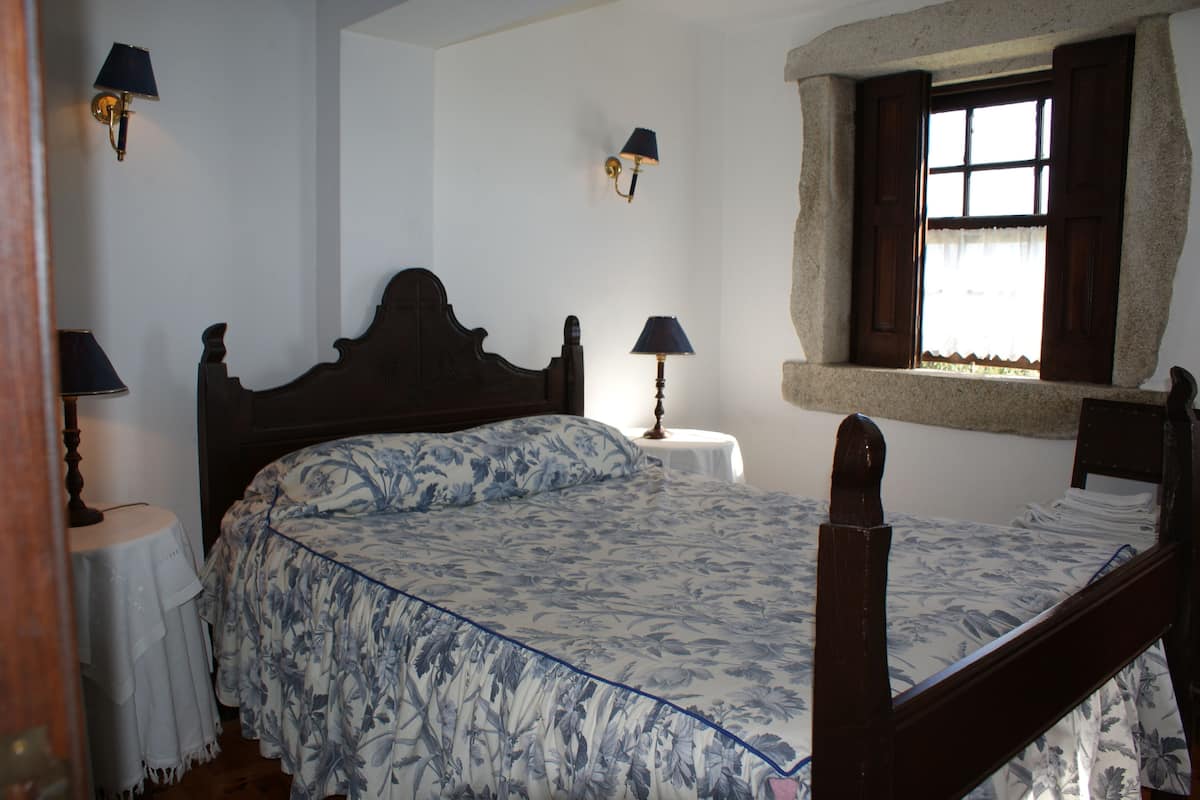 A bedroom with a wooden bed, two bedside lamps, a small round table, and a window with curtains.