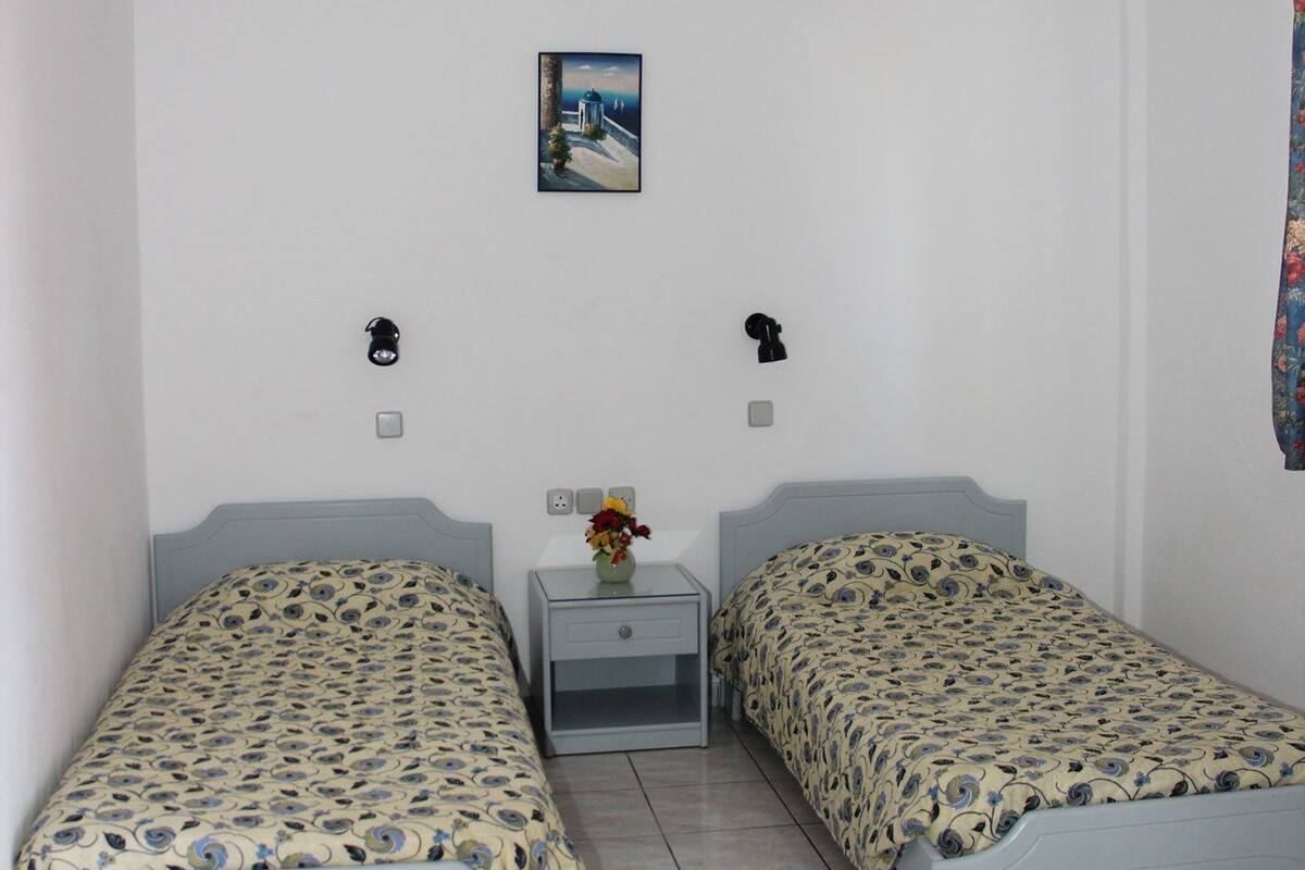 A bedroom with two single beds, a nightstand, a wall-mounted lamp, a small painting, and a curtain.