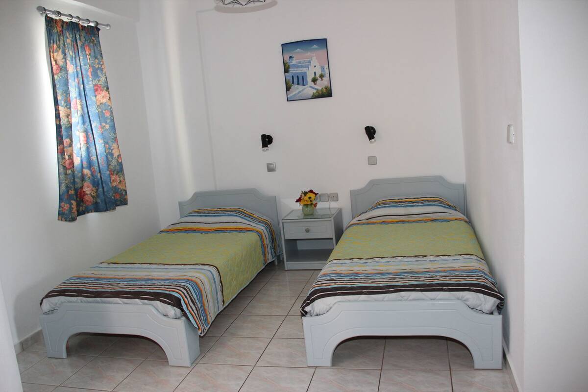 A small, clean room with two single beds, a nightstand with a vase, and a wall-mounted picture.