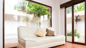 A white sofa with two pillows in a room with large windows and a view of greenery outside. - Rasta Hotel (Koh Samui)
