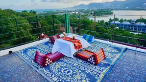 A rooftop terrace with a table set for dining, surrounded by patterned cushions and a view of a lake and mountains. - Rasta Hotel (Koh Samui)