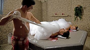 A man applying foam to a couple in a tiled room with a brick wall and a hanging plant.