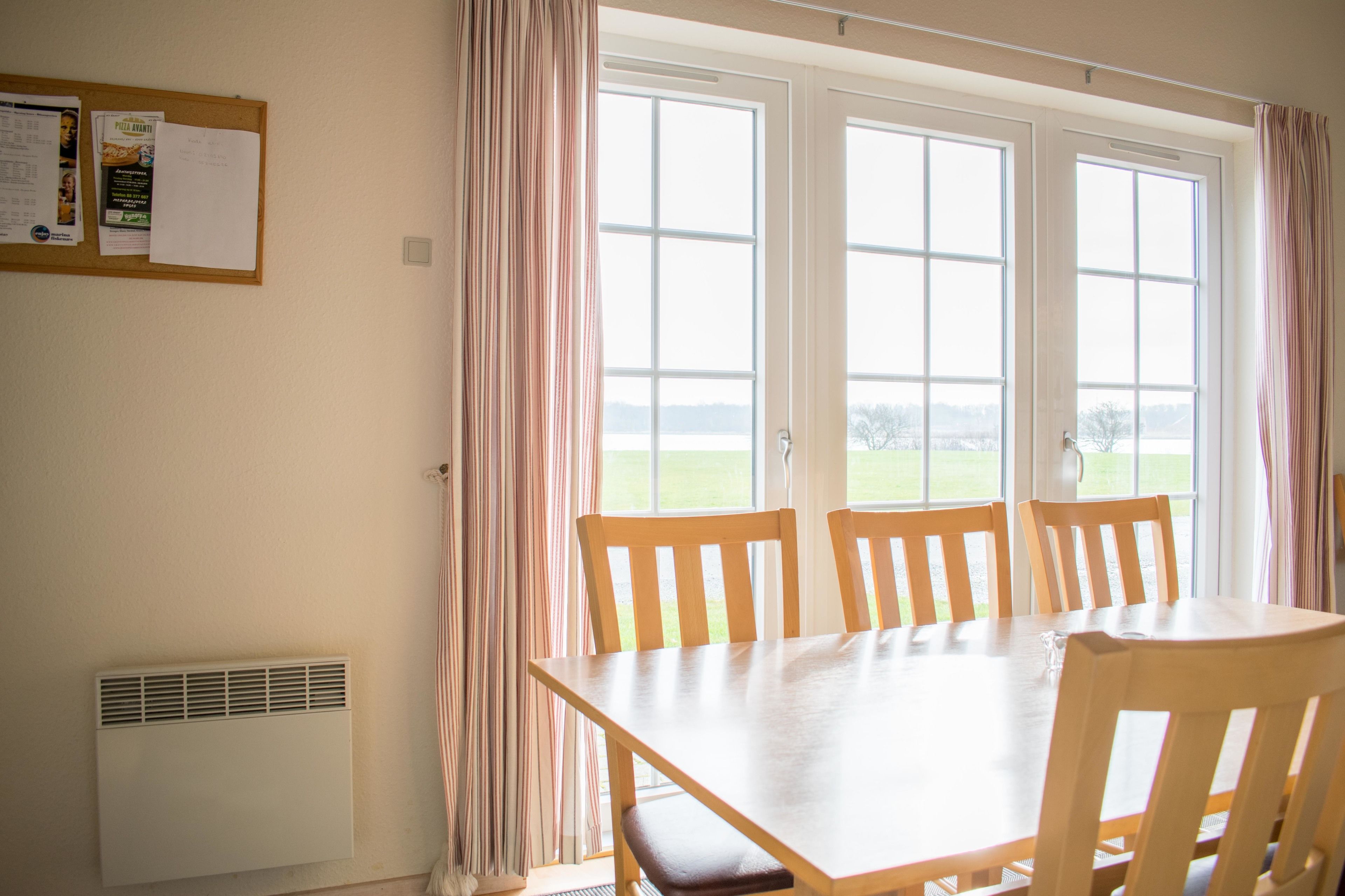 A dining area with a table and chairs, a bulletin board with notices, and a view through large windows.