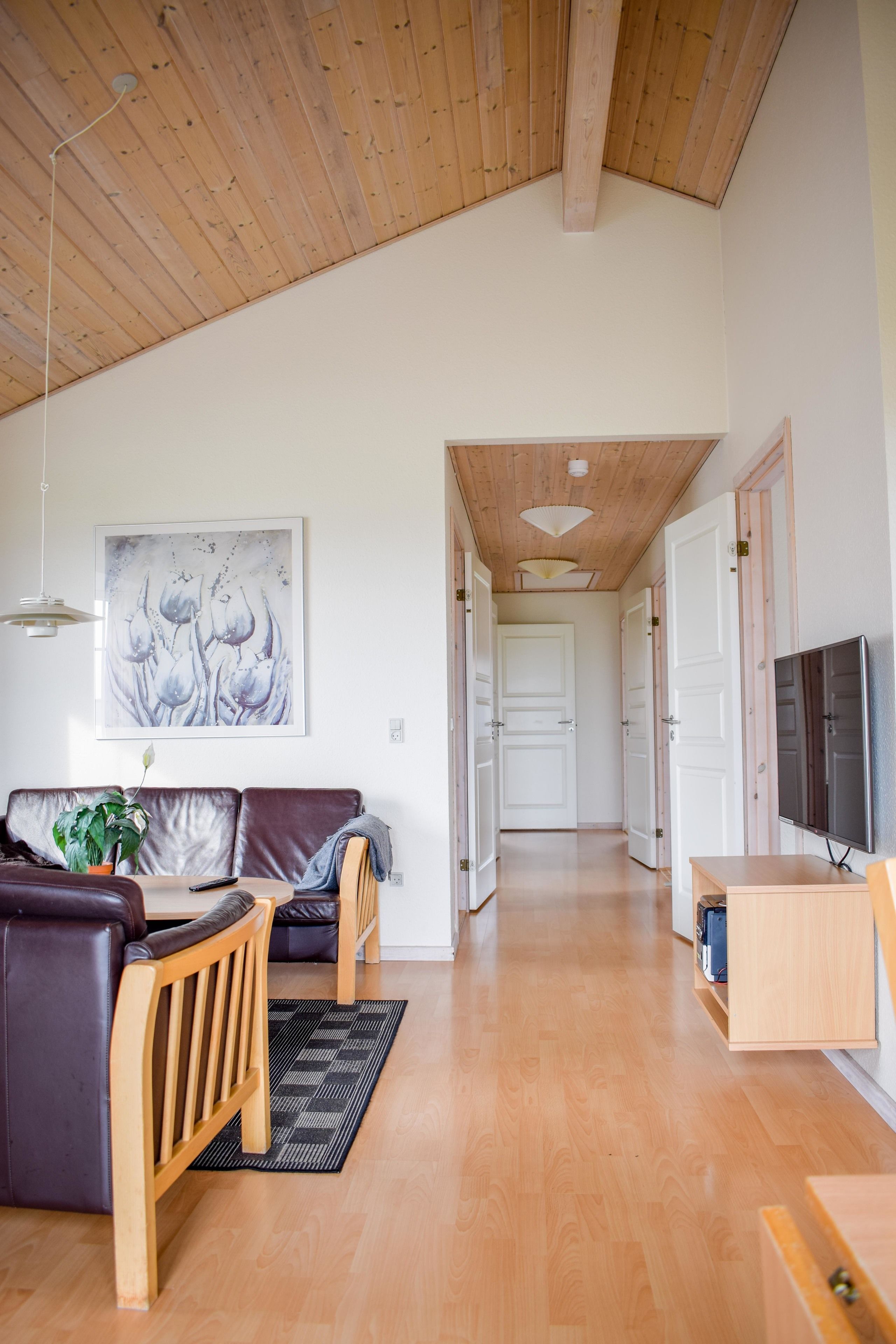 Een ruime woonkamer met een houten plafond, een bank, een salontafel, een televisie en een deur naar een andere kamer.