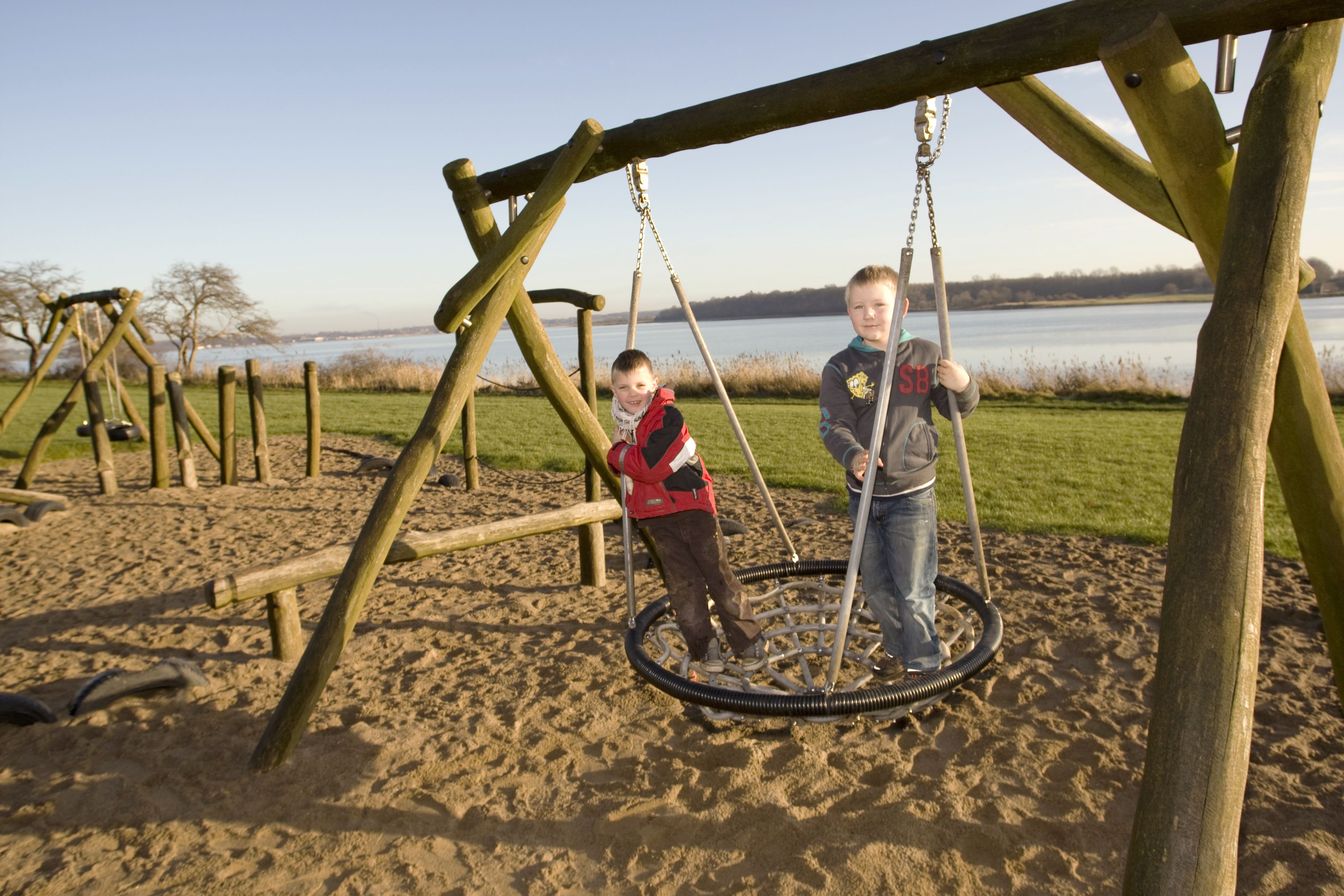 A wooden swing set with a tire swing, located on a sandy area near a body of water.