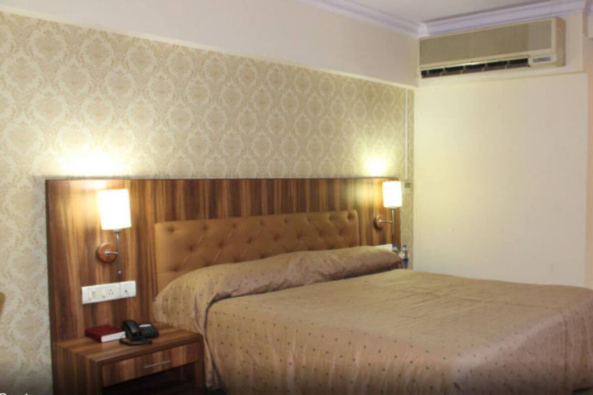 A hotel room with a large bed, two bedside lamps, a wooden nightstand with a telephone, and an air conditioning unit above the bed.