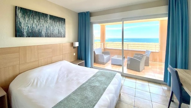 A hotel room with a large bed, a view of the ocean, and a balcony with outdoor seating.