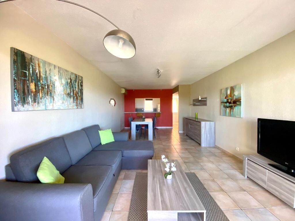 Een moderne woonkamer met een grijze bank, een salontafel en een flatscreen televisie. Op de achtergrond is een keuken met rode muren en een eettafel te zien.
