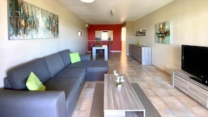 A modern living room with a grey sofa, a coffee table, and a flat-screen TV. There is a kitchen area in the background with red walls and a dining table.
