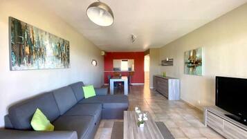 Een moderne woonkamer met een grijze bank, een salontafel en een flatscreen televisie. Op de achtergrond is een keuken met rode muren en een eettafel te zien.