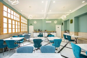 Een eetruimte met teal stoelen en witte tafels, grote ramen en een keuken op de achtergrond.