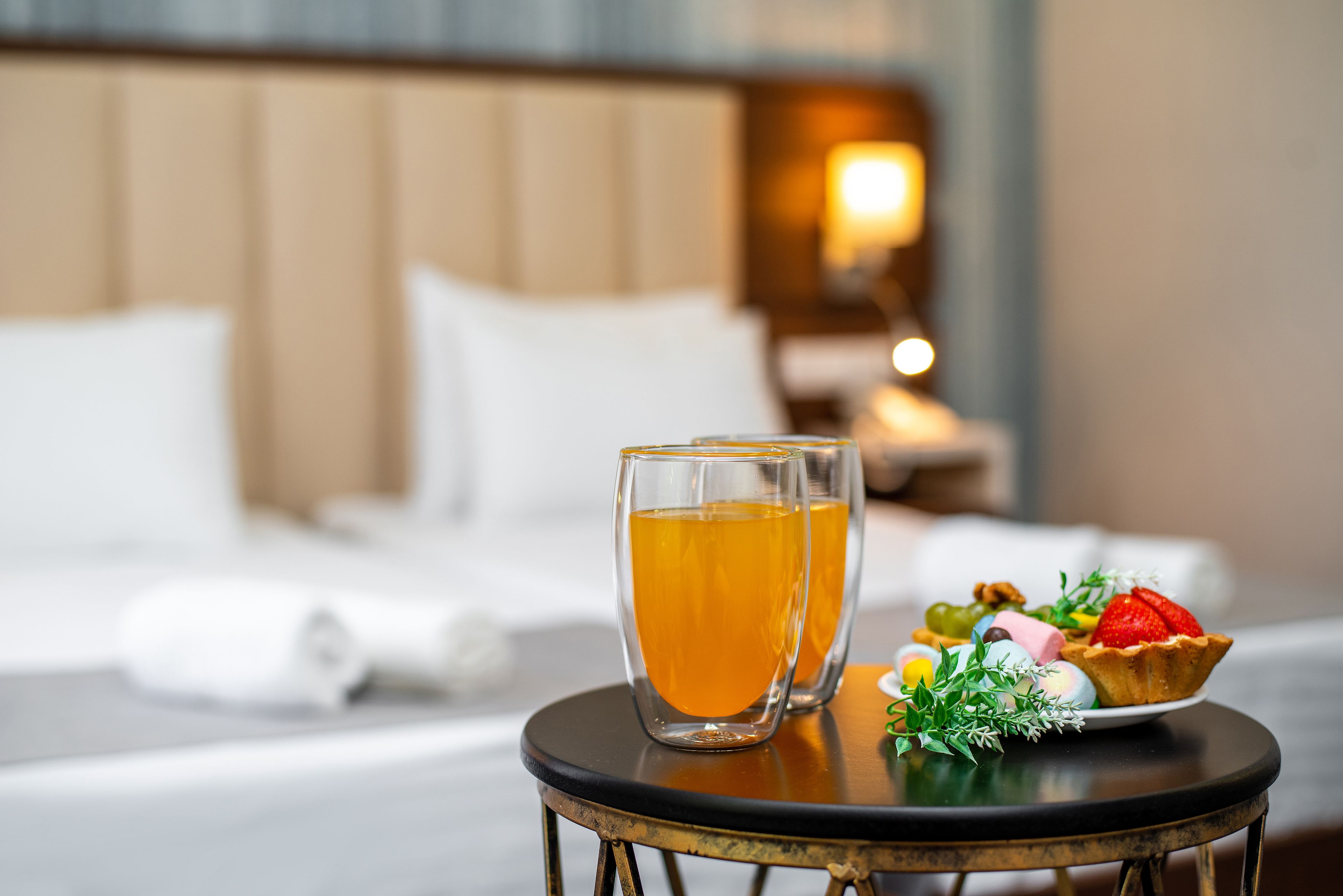 A neatly made bed with white linens, a small round table with a glass of amber liquid and a dessert, and a nightstand with a lamp in a hotel room.