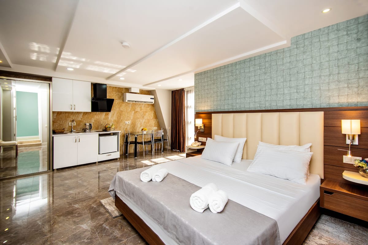 A modern hotel room with a large bed, a separate dining area, and a kitchenette.