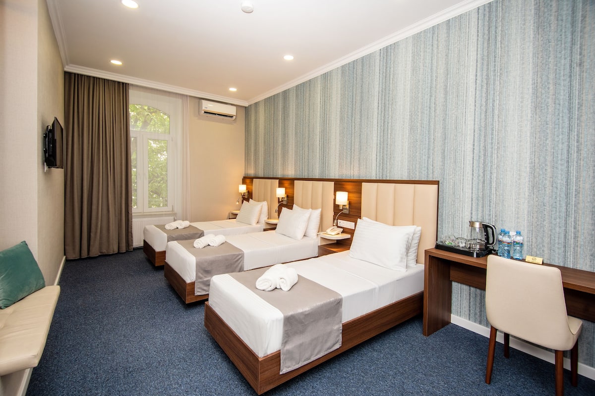 A hotel room with three single beds, a desk with a chair, a sofa, and a window with curtains.