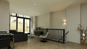Un espacio habitable amplio con suelo de diseño herringbone, ventana de marco negro, sofá negro, sillón gris, una mesita y un jarrón decorativo.