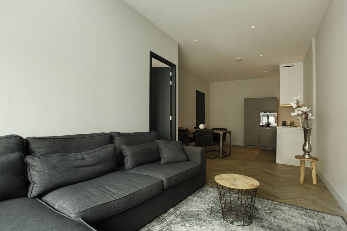 Un salon moderne avec un canapé gris, une table basse ronde et un petit tabouret en bois. On aperçoit à l’arrière-plan un coin cuisine avec une table à manger et des chaises.