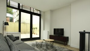Una sala moderna con un sofá seccional gris, dos mesas redondas de madera, un mueble de TV negro y un ventanal con vistas a un edificio y un árbol.