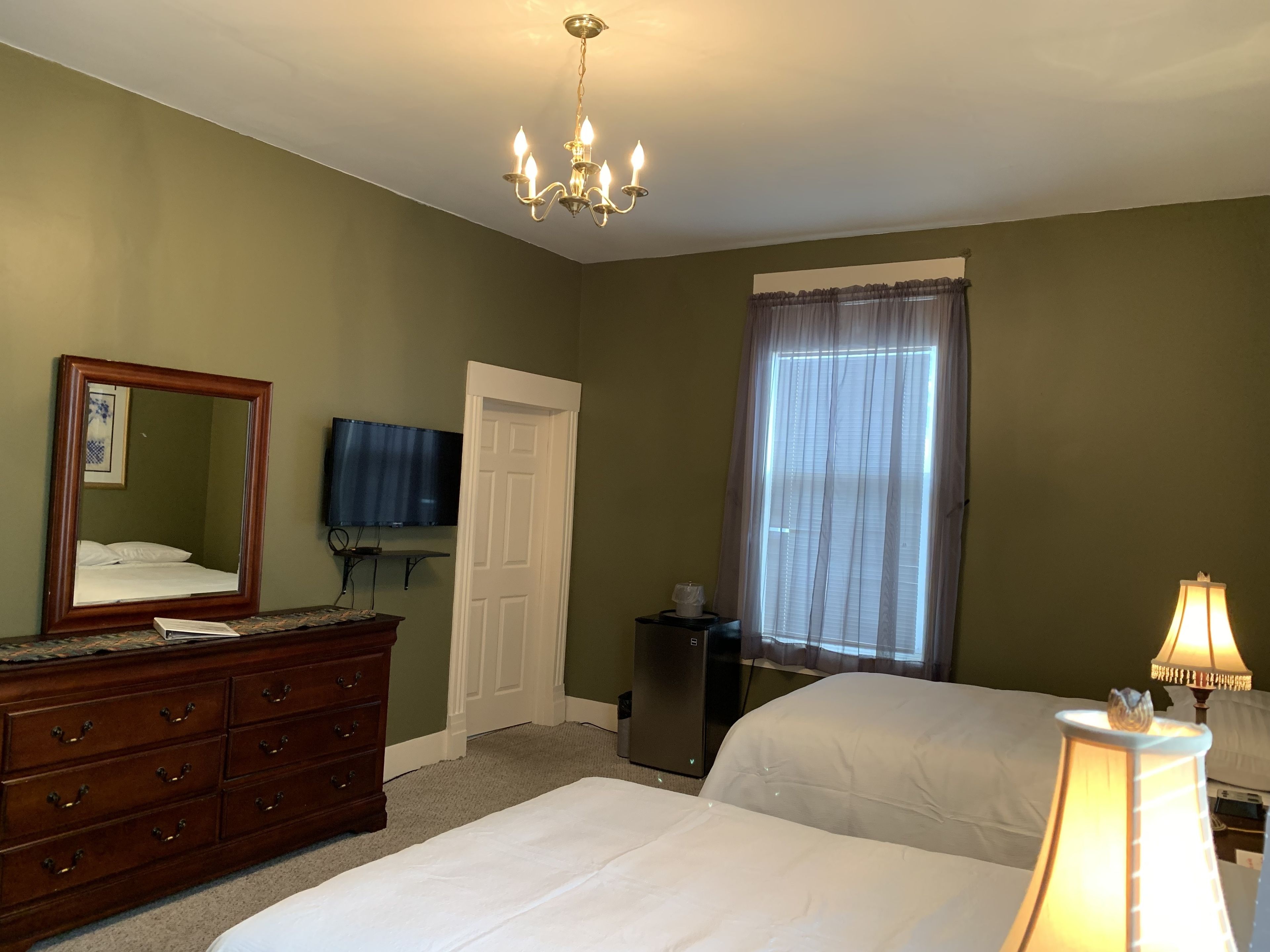 A hotel room with a large bed, a dresser, a television, a chandelier, and a window with curtains.