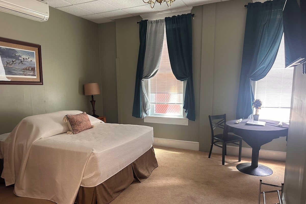 A hotel room with a bed, a desk, a chair, a lamp, and a window with curtains.