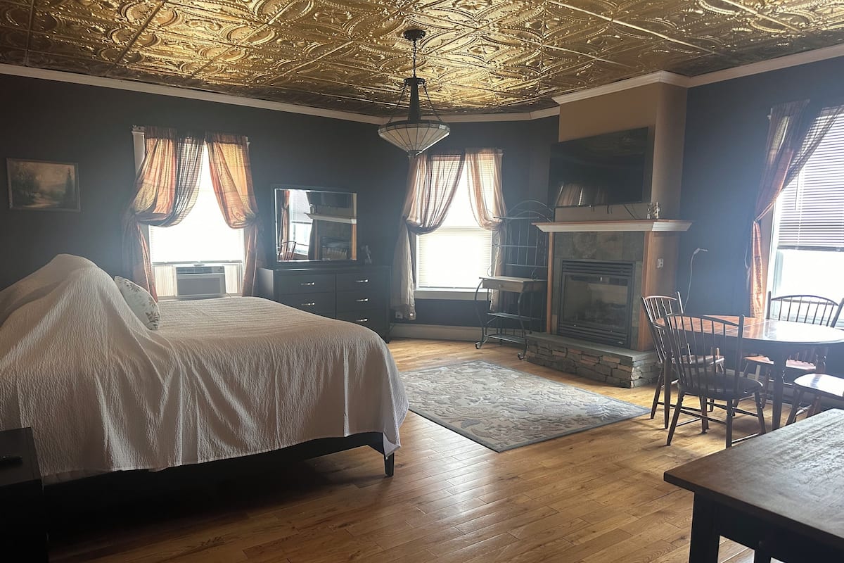 A bedroom with a bed, a dresser, a mirror, a fireplace, and a dining area with chairs and a table.