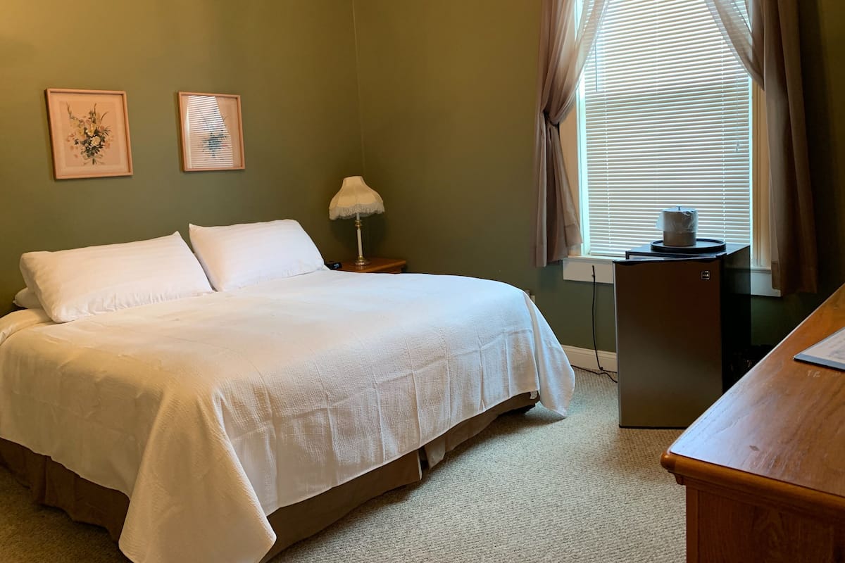 A bedroom with a bed, a nightstand with a lamp, a dresser, and a window with blinds.