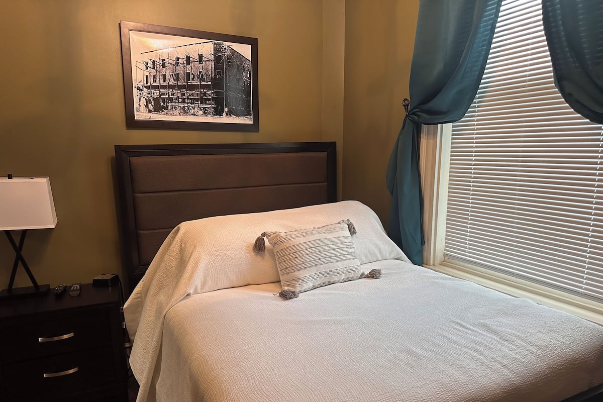 A neatly made bed with a pillow, a framed picture on the wall, a bedside lamp, and a window with curtains.
