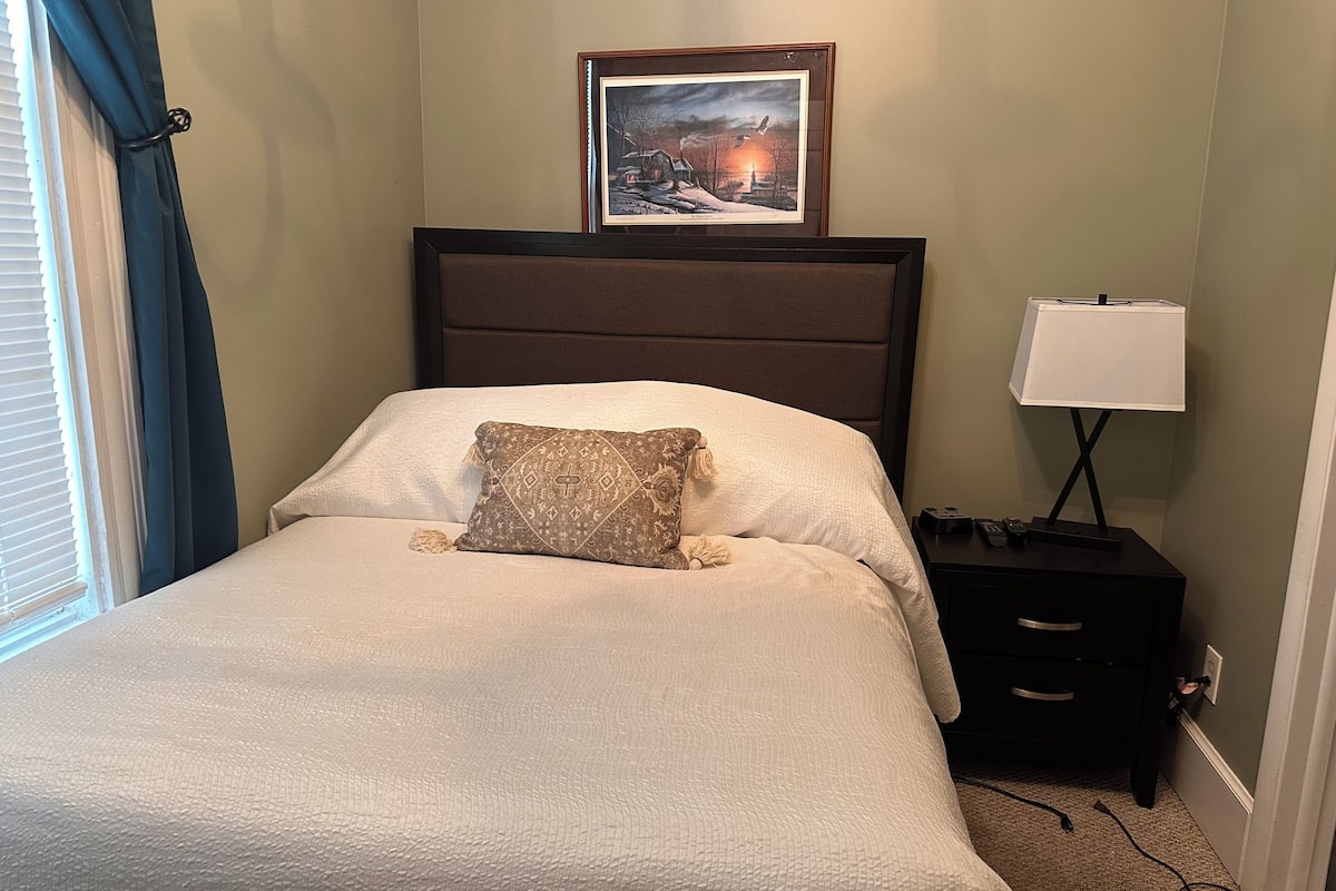 A neatly made bed with a patterned pillow, a framed picture on the wall, a bedside lamp, and a small bedside table with a remote control.
