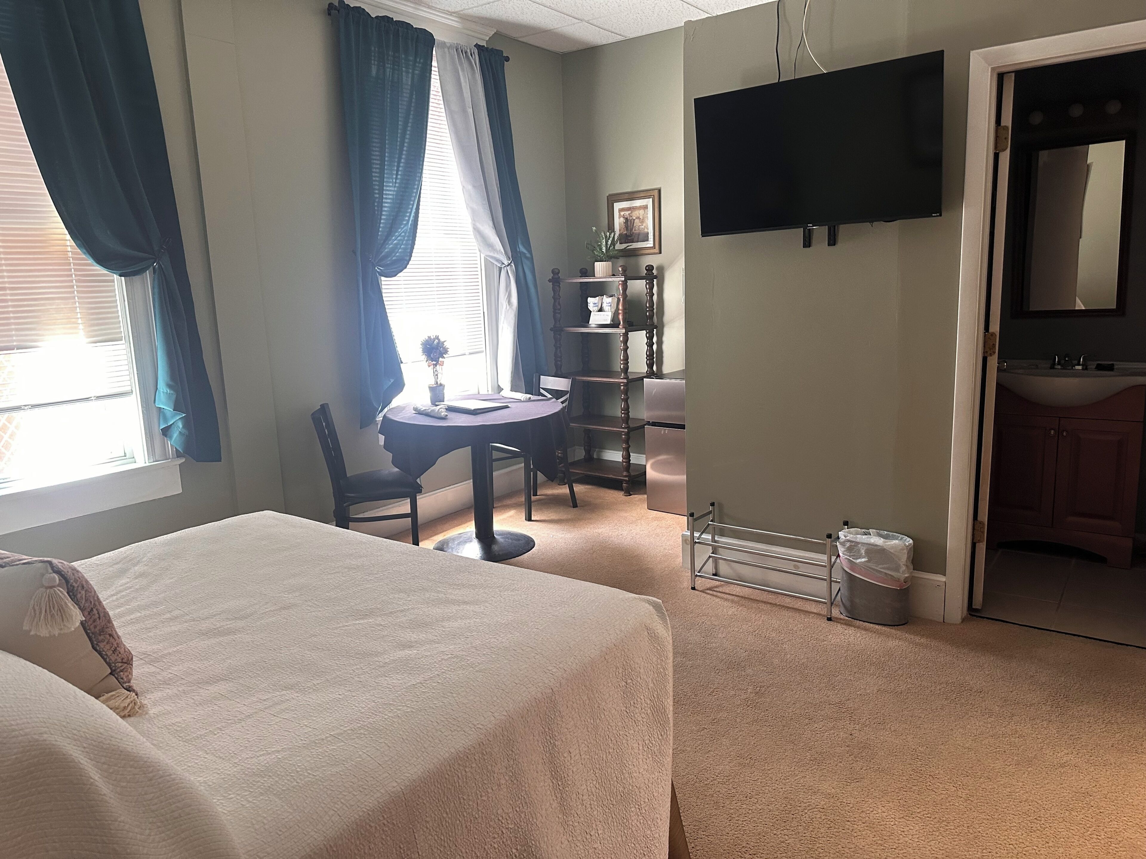 Habitación de hotel con cama, una mesa de comedor con sillas, televisor, pequeña cocina y ventana con cortinas.