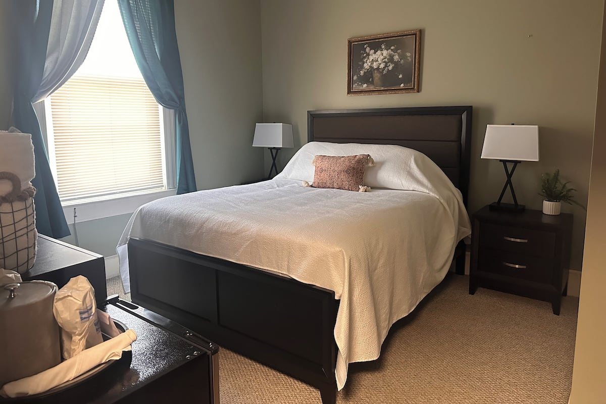 A neatly made bed with a pillow, two bedside lamps, a nightstand with a plant, and a framed picture on the wall.