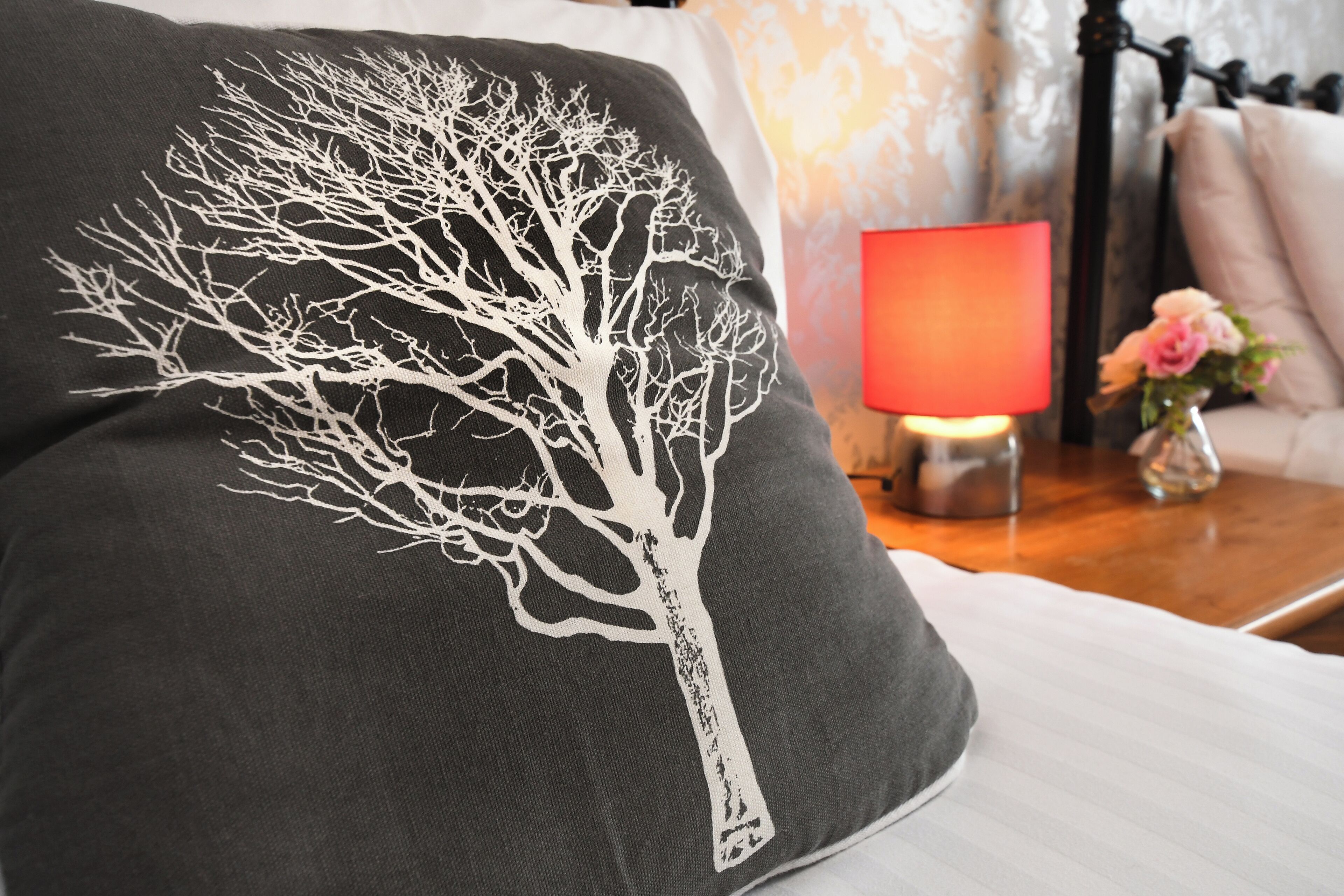 A pillow with a white tree design, a bedside lamp, a vase with flowers, and a bed with white bedding.