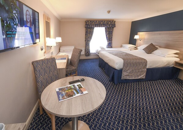 A hotel room with a bed, a desk, a chair, a TV, and a round table with a magazine.