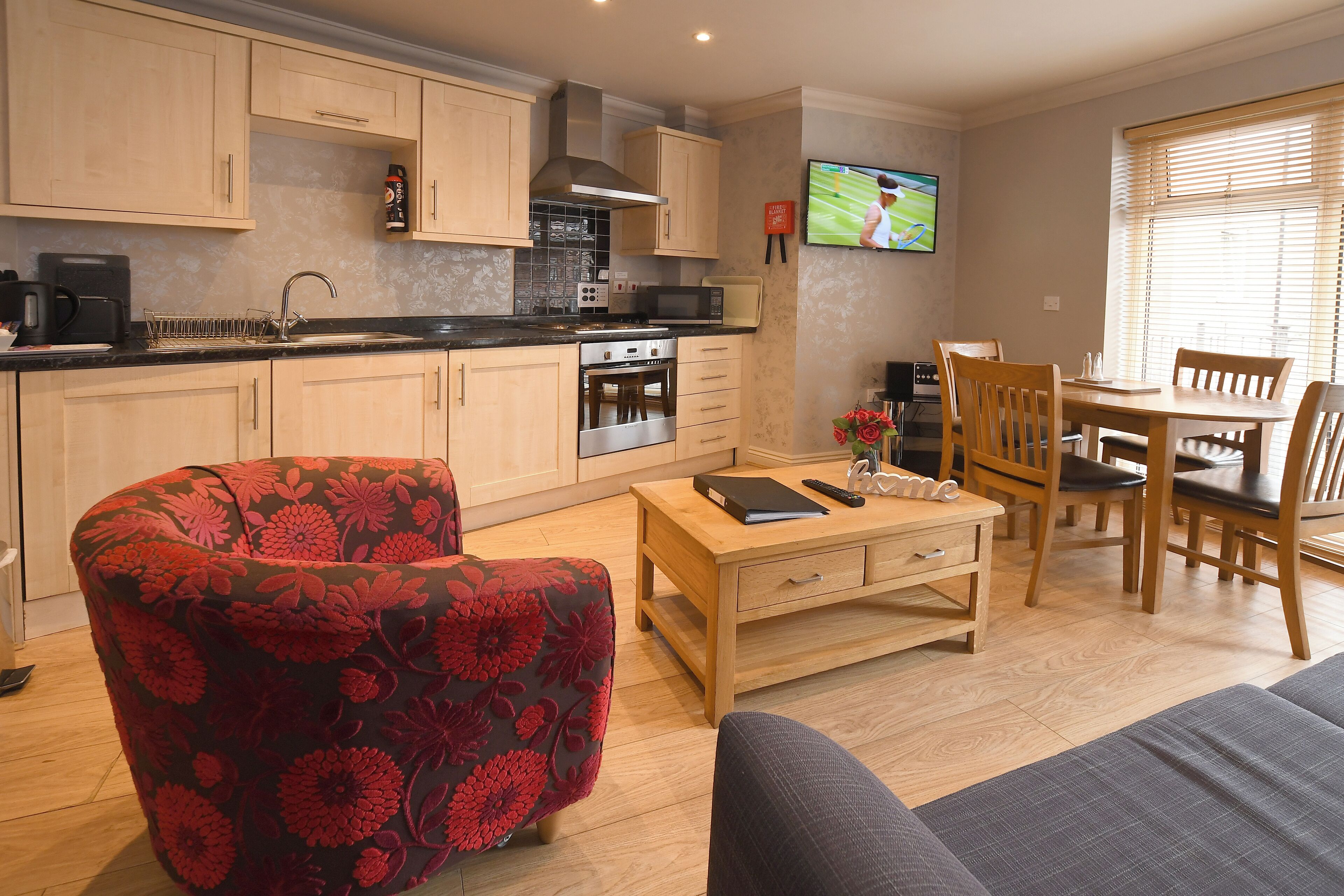 A modern kitchen with wooden cabinets, a microwave, and a stove. There is a dining area with a table and chairs, and a sofa in the living room.