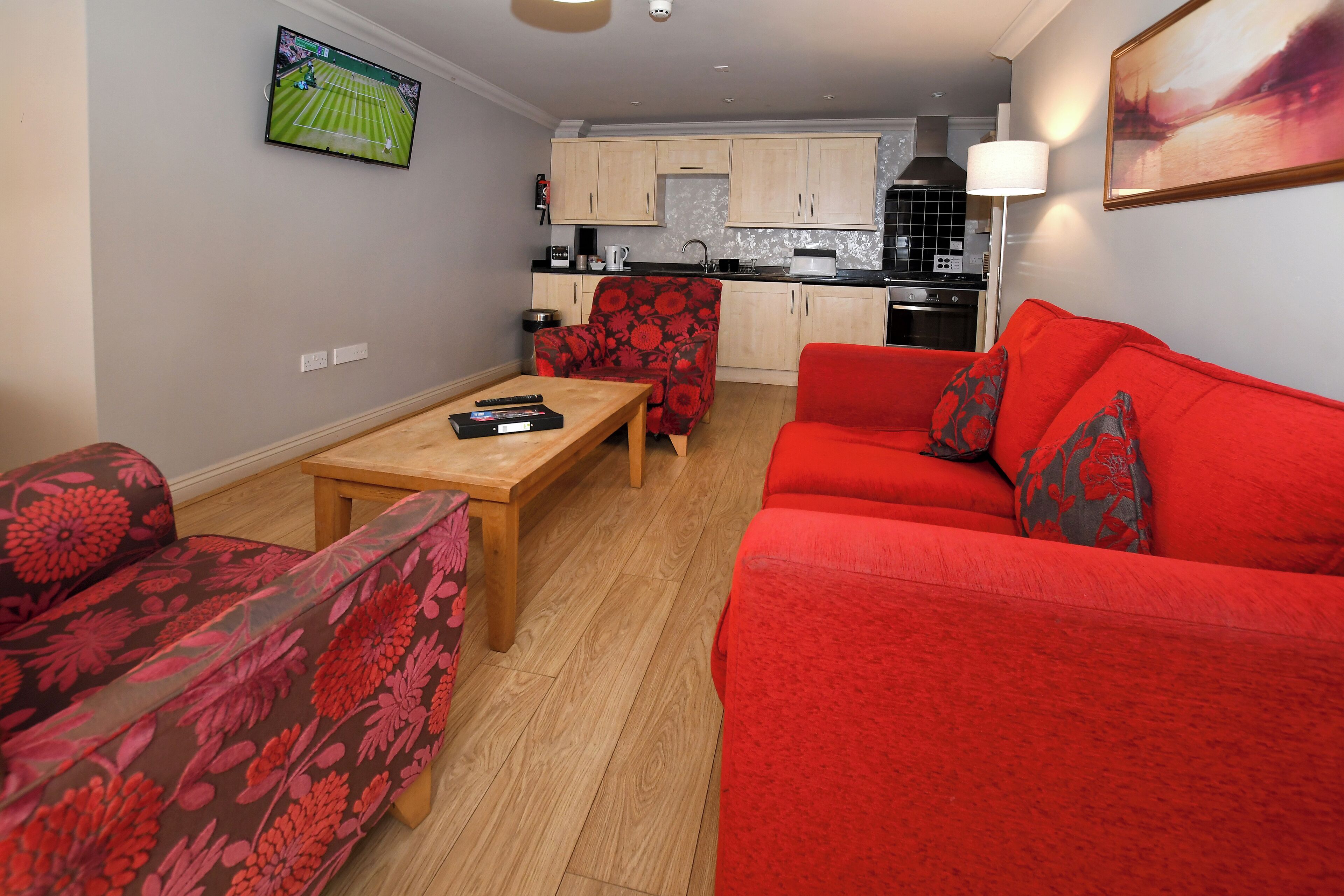 Una sala de estar con un sofá rojo, un sillón con estampado floral y una mesa de centro de madera. Se ve una cocina al fondo, con armarios de tonos claros y una estufa.