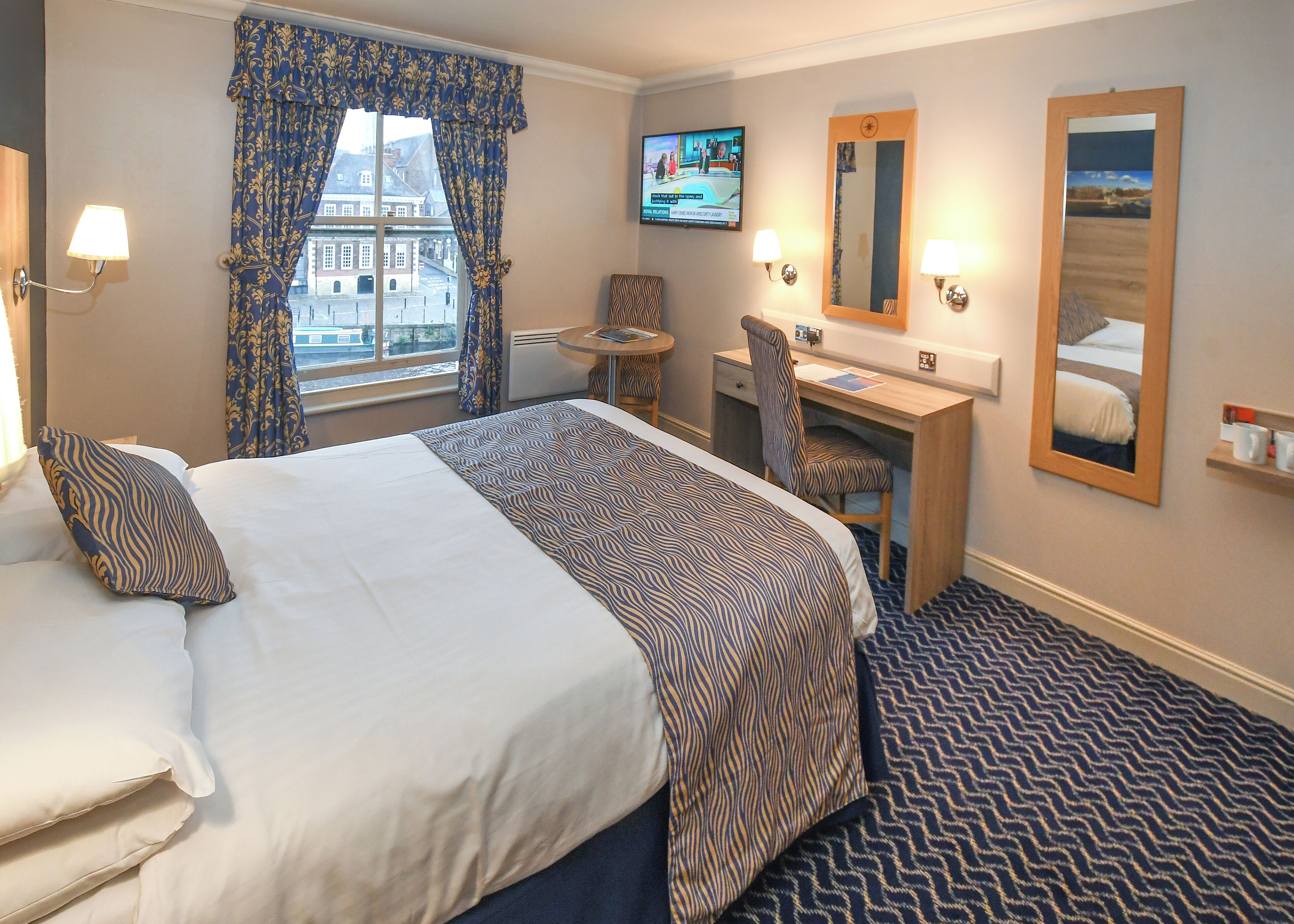 A hotel room with a large bed, a desk, a chair, a TV, and a view of the city through the window.