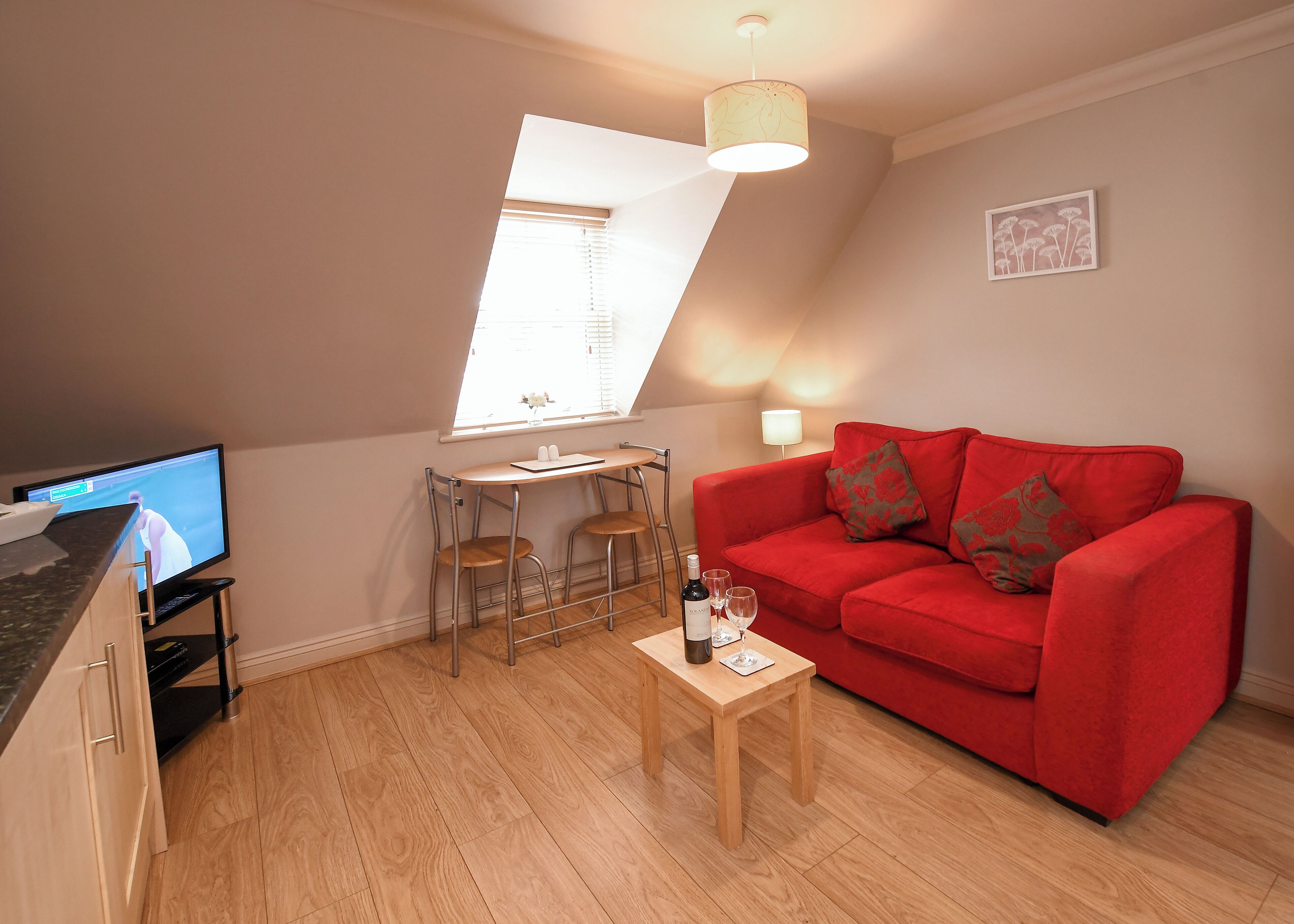 Un salon chaleureux avec un canapé rouge, une petite table en bois sur laquelle se trouvent une bouteille et des verres, un téléviseur posé sur un meuble et une fenêtre avec des stores.