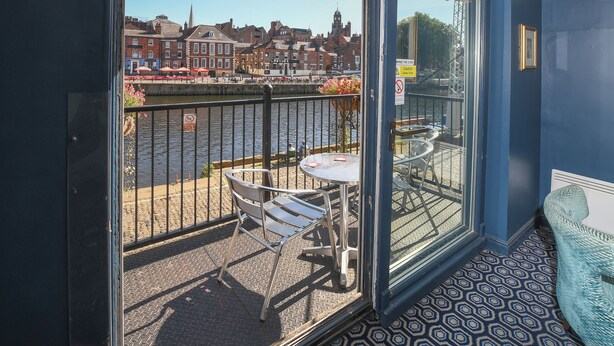 Un balcón con una mesa y sillas, vista a un río y edificios al fondo.