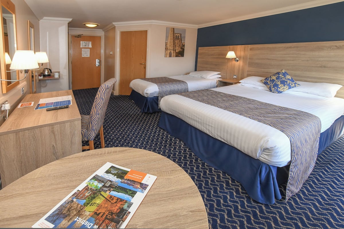 Habitación de hotel con dos camas, un escritorio con silla, una mesa redonda con un folleto y una lámpara de pared.