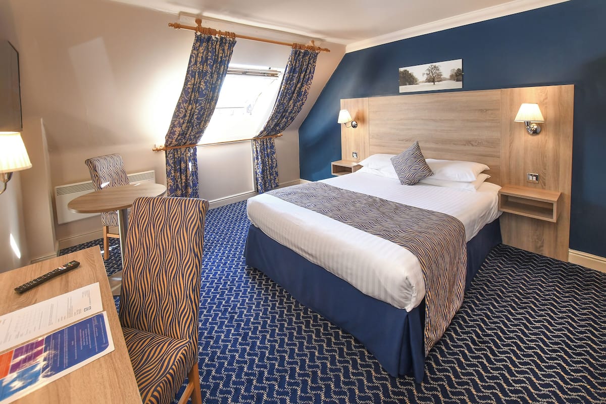 Habitación de hotel con una cama grande, un escritorio con silla, una mesita pequeña y una ventana con cortinas.