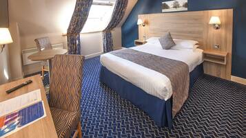 A hotel room with a large bed, a desk with a chair, a small table, and a window with curtains.