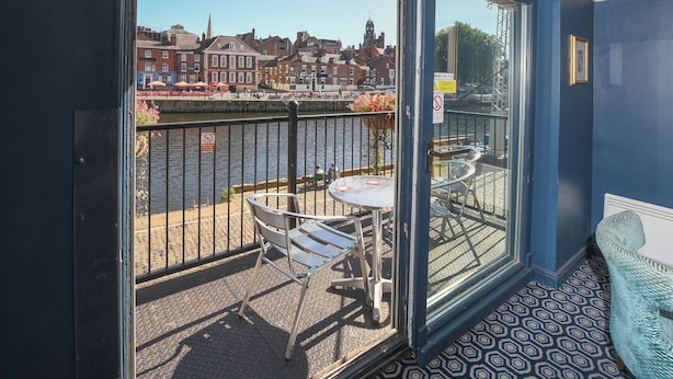 Un balcón con una mesa y sillas, vista a un río y edificios al fondo.