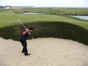 A person playing golf on a course with sand bunkers, water hazards, and a clubhouse in the background.